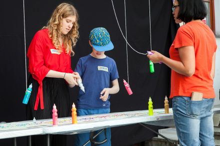 Chłopiec maluje swój obrazek farbami, które wiszą nad stołem. Pomagają mu dwie prowadzące.