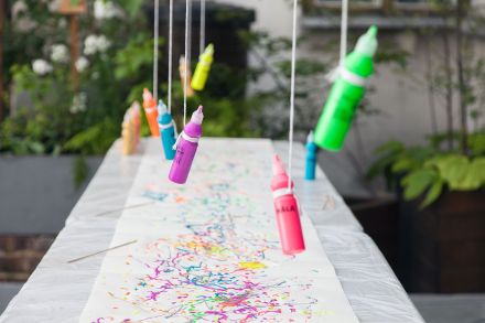 Długi stół  osłonięty folią. Na nim leży karton wielkości całego stołu. Na sznurkach wiszą kolorowe farby, które kapiąc na kartkę, tworzą obrazek.