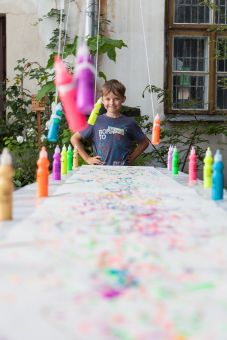 Zbliżenie na fragment stołu i kartki, na której powstały kolorowe mazaje. Dookoła kartki na stole stoją farby, a na końcu stołu stoi chłopiec i się uśmiecha.