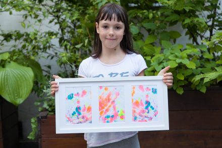 Dziewczynka prezentuje skończony obrazek. Za nią widać wysokie krzewy.