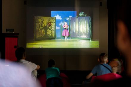 Zbliżenie na scenę, gdzie wystawiany jest spektakl. Na scenie aktorka w różowej sukience. Dekoracja to las z papieru.