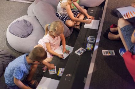 Zdjęcie z lotu ptaka. Dzieci siedzą na podłodze. Na podwyższeniu leżą kartki. Dzieci rysują.