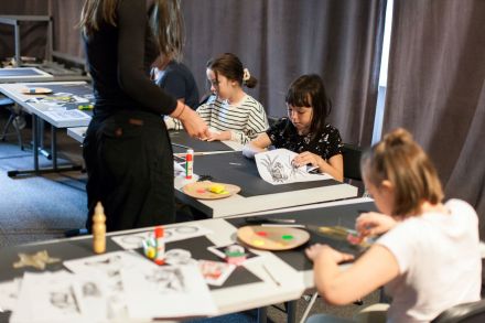 Dzieci przy stole rysują farbami. Stoi przy nich prowadząca warsztaty.