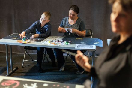 Zbliżenie na chłopca, który sięga ręką do farb praz na panią, która wycina coś z papieru nożyczkami.