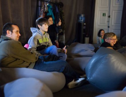 Zbliżenie na publiczność. Ludzie siedzą lub leżą na pufach-workach.