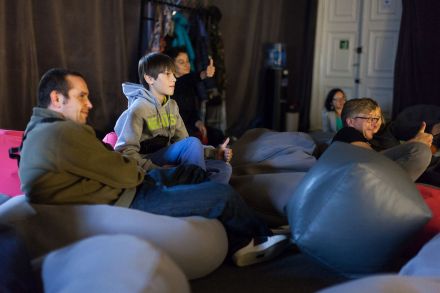 Zbliżenie na publiczność. Ludzie siedzą lub leżą na pufach-workach.