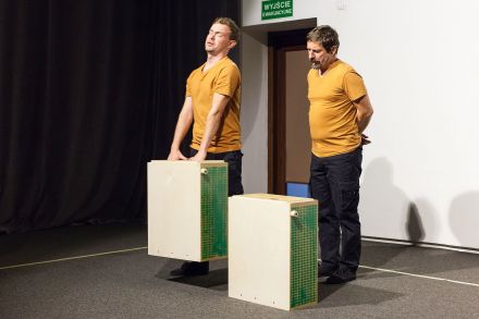 Dwaj aktorzy w żółtych koszulkach. Jeden dźwiga ciężką torbę. Drugi już ją postawił na ziemi.