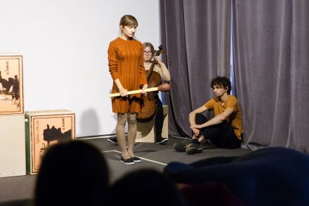 Scena. Kobieta gra na  na wiolonczeli, mężczyzna siedzi oparty o kotarę sceny. Przed nimi stoi kobieta z wielką zapałką w dłoni.