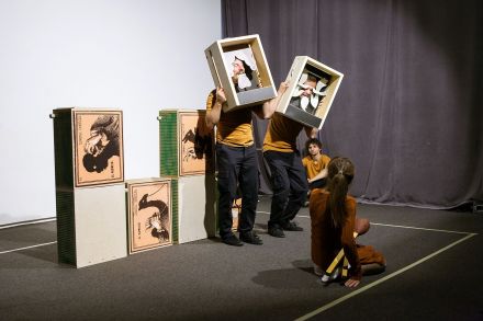 Scena. Dwaj aktorzy z drewnianymi skrzynkami na głowach oraz siedząca przed nimi kobieta.