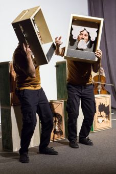 Dwaj aktorzy z drewnianymi skrzynkami na głowach. Mają papierowe brody i długie włosy.