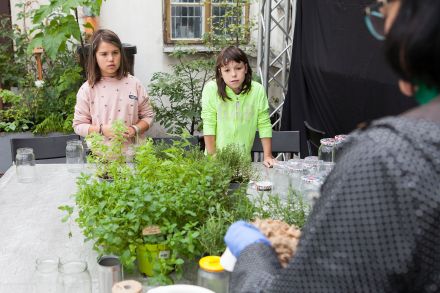 Stół, na którym stoją kwiaty w doniczce. Po jednej stronie stoją dwie dziewczynki, po drugiej stronie prowadząca wyjaśnia co należy zrobić.