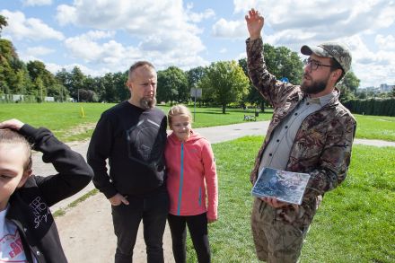Przewodnik pokazuje inne zdjęcie. Unosi rękę ku górze. Uczestnicy spaceru mu się przyglądają.