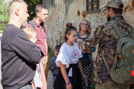 Przewodnik i uczestnicy spaceru na tle starego zniszczonego budynku.