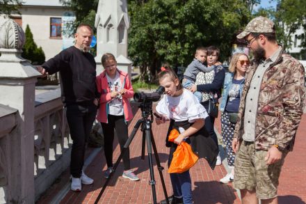 Dziewczynka patrzy przez lunetę. Dookoła niej stoją przewodnik i reszta uczestników spaceru.