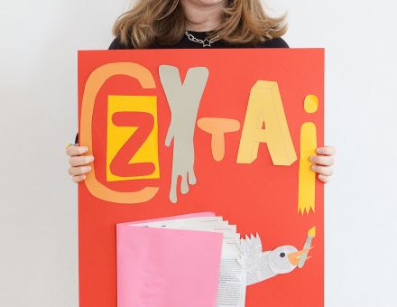 Dziewczynka trzyma w ręce samodzielnie wykonany plakat z napisem "Czytaj" oraz książką.
