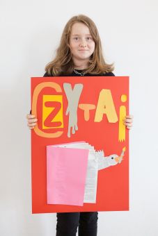 Dziewczynka trzyma w ręce samodzielnie wykonany plakat z napisem "Czytaj" oraz książką.