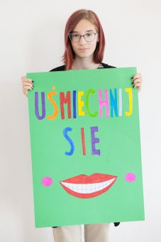 Dziewczyna trzyma w ręce samodzielnie wykonany plakat z napisem "uśmiechnij się" i ustami.