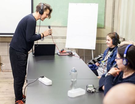 Prowadząca mówi do mikrofonu. Siedząca naprzeciw niej dziewczynka ma nałożone  słuchawki i się uśmiecha.
