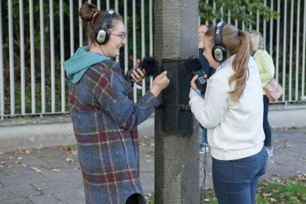 Dwie dziewczyny przykładają mikrofon do betonowego słupa i słuchają jego odgłosów.