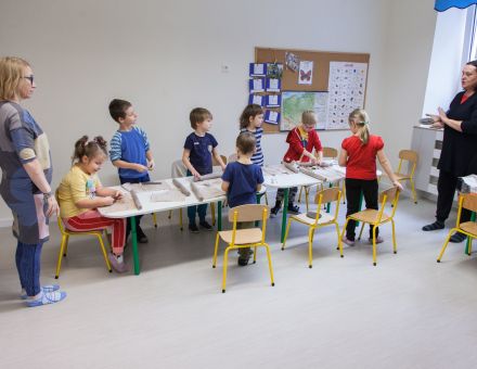 Dzieci siedzą przy stoliku. Dwie prowadzące stoją i je instruują.