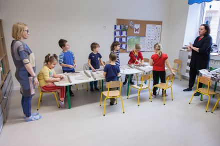 Dzieci siedzą przy stoliku. Dwie prowadzące stoją i je instruują.