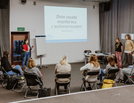 Uczestnicy siedzą w półkolu przed ścianą, na której wyświetlana jest prezentacja pod hasłem "Złote zasady współpracy z wolontariuszami".