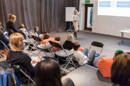 Na pierwszym planie uczestnicy spotkania. W tle prowadząca, która tłumaczy zagadnienia za pośrednictwem prezentacji multimedialnej.