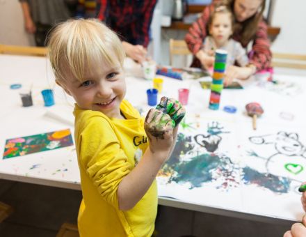 Dziewczynka pokazuje ręce zabrudzone farbą i uśmiecha się do kamery.