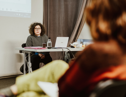 Prowadząca spotkanie siedzi przy biurku. Za nią na ścianie wyświetlana jest prezentacja multimedialna.  przed nią siedzą uczestnicy.