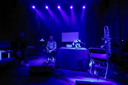 Na scenie po lewej stronie wokalista, na środku tańczący robot, a po prawej stronie saksofonista.