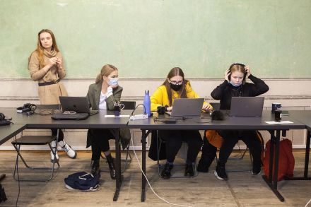 Trzy dziewczyny siedzą przy stole i pracują na laptopach. Obok oparta o ścianę jest czwarta kobieta.