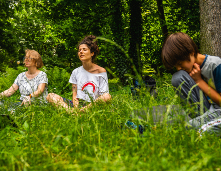 Dwie kobiety medytują na trawie. Obok siedzi chłopiec i patrzy w trawę.