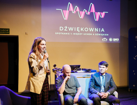 Kobieta stoi przy stoliku, a dwaj mężczyźni się jej przysłuchują. Na ścianie wyświetlone jest logo Dźwiękowni.