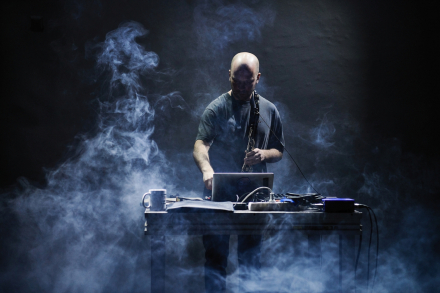 Muzyk stoi przy konsoli. W ręce trzyma saksofon. Scena jest zadymiona,