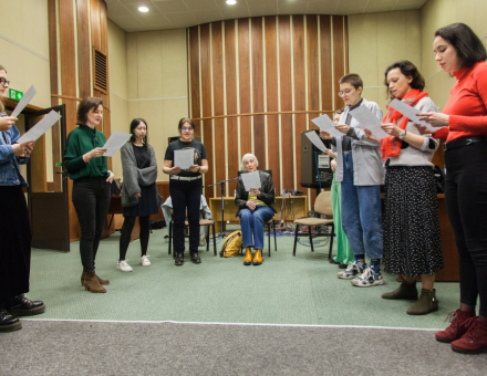 Grupa dziewięciu osób stoi w półokręgu. Każdy trzyma w ręce kartkę i czyta tekst.