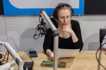 Dziewczyna siedzi przy mikrofonie dla spikerów radiowych i czyta tekst z kartki.