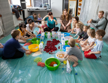 Uczestnicy warsztatu wraz z rodzicami i prowadzącą siedzą na podłodze w kole. Pośrodku stoją miski, butelki, mąka, galaretka i inne produkty, którymi będą bawiły się dzieci.