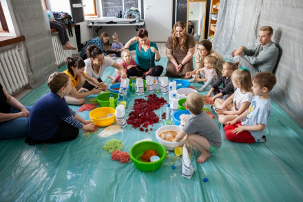 Uczestnicy warsztatu wraz z rodzicami i prowadzącą siedzą na podłodze w kole. Pośrodku stoją miski, butelki, mąka, galaretka i inne produkty, którymi będą bawiły się dzieci.