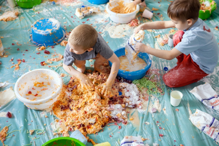 Chłopiec bawi się kolorową masą, którą wysypał z miski. Obok niego drugi malec wsypuje do miski mąkę.