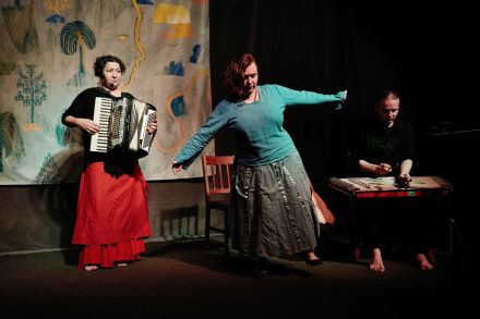 Zdjęcie trzech osób z zespołu, z czego kobieta po środku tańczy, a dwie osoby po bokach grają na  swoich instrumentach.