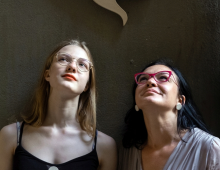 Dwie kobiety patrzą w niebo. Nad ich głowami wisi tabliczka z napisem "głowa do góry".