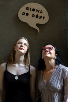 Dwie kobiety patrzą w niebo. Nad ich głowami wisi tabliczka z napisem "głowa do góry".