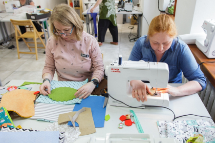 Dwie kobiety siedzą przy stole. Jedna szyje na maszynie, a druga odrysowuje kwiatka na na materiale.