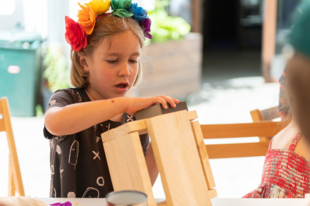 Dziewczynka z wiankiem na głowie trzyma w ręce drewniane elementy do budowy domu dla pszczół.