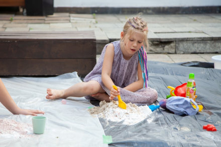 Dziewczynka siedzi na czarnej płachcie wyłożonej na ziemi i bawi się piaskiem.