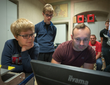 Mężczyzna obsługuje komputer. Dookoła niego stoją uczestnicy warsztatów i wpatrują się, co się dzieje na ekranie.