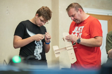 Mężczyzna trzyma w ręce samolot. Obok chłopiec trzyma ostatnie elementy do skrzydeł maszyny.