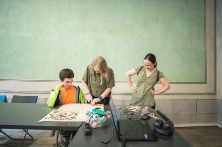 Kobieta wycina gotowy projekt z płyty. Przygląda jej się chłopiec i druga kobieta.