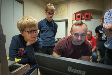 Mężczyzna obsługuje komputer. Dookoła niego stoją uczestnicy warsztatów i wpatrują się, co się dzieje na ekranie.