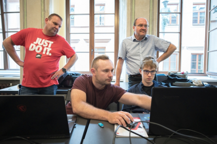 Mężczyzna obsługuje komputer. Obok niego siedzi chłopiec. Z tyłu stoi dwóch mężczyzn.